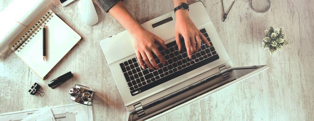 Blog image - looking down on a person typing on a laptop with a pen and paper next to them